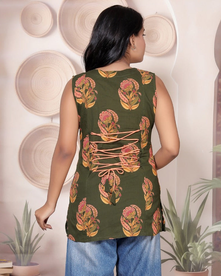 A woman shown from the back wearing a sleeveless olive green tunic top with a pink and orange floral pattern and blue wide-leg jeans, standing in a styled room with plants and arches.