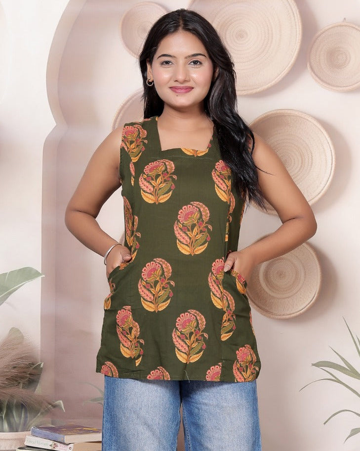A woman modeling a sleeveless olive green top with a yellow and orange floral pattern, paired with wide-leg blue jeans, standing in a styled indoor setting with plants and decorative baskets.