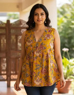 Woman wearing a yellow floral sleeveless top and blue jeans standing on a tiled floor with plants in the background.